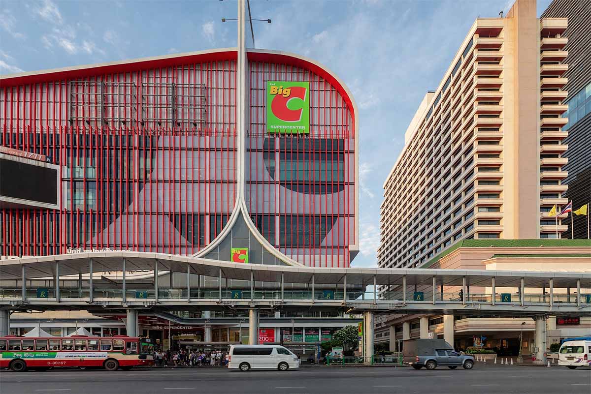 Fachada exterior del centro comercial Big C Supercenter Ratchadamri en Bangkok, Tailandia, con su distintivo edificio rojo, un puente peatonal elevado enfrente y tráfico urbano en primer plano.